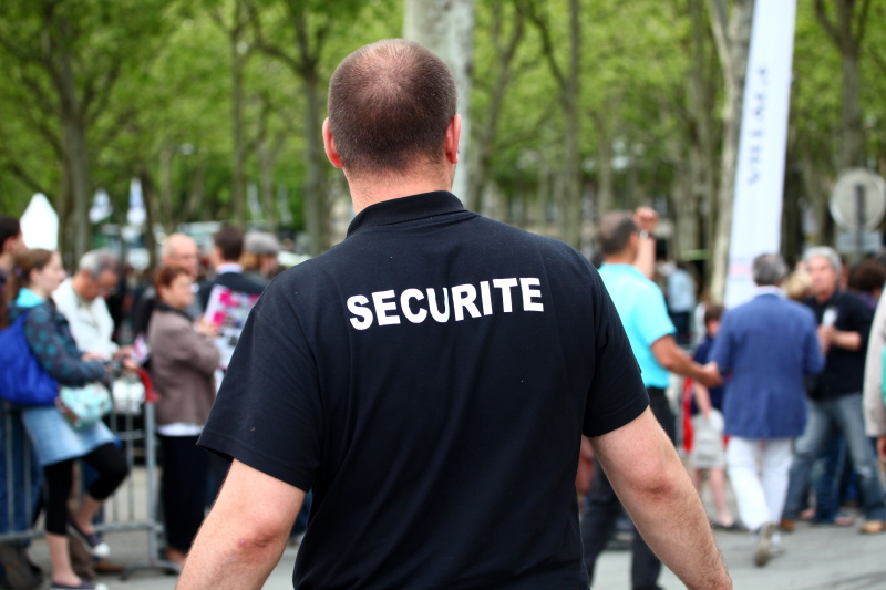 Vue de dos sur un agent de sécurité privée lors d'un événement public, avec de la foule au second plan.